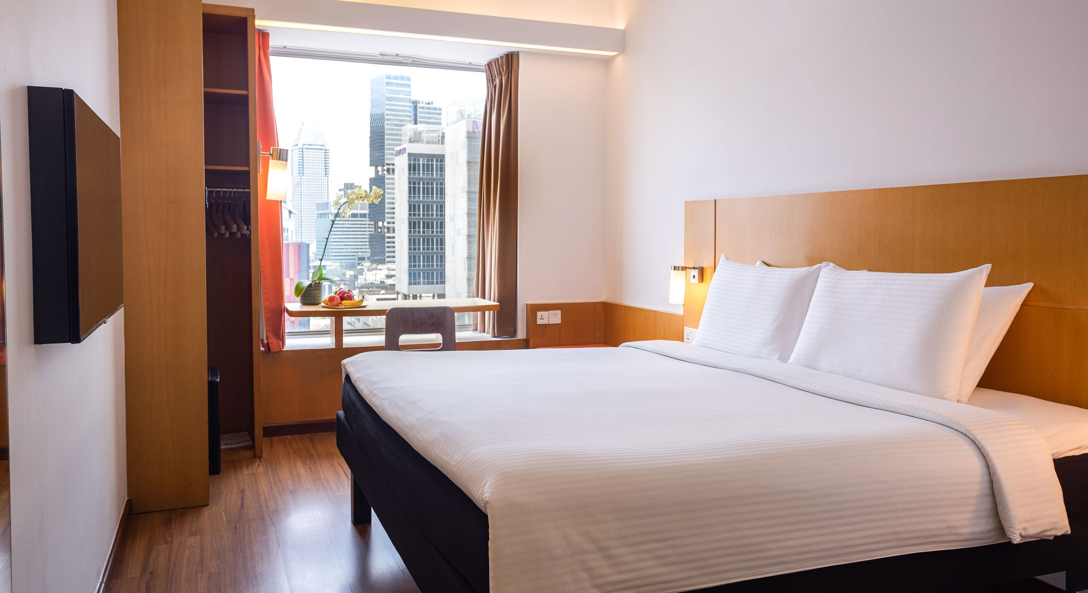 Modern hotel room with a king-size bed, a large window offering a city view, a flat-screen TV, and a desk.