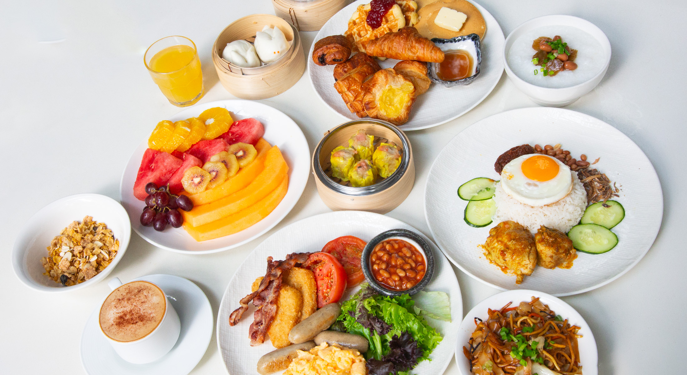 A variety of international and local breakfast items, including fruits, pastries, eggs, beans, noodles, and beverages, served on white plates.