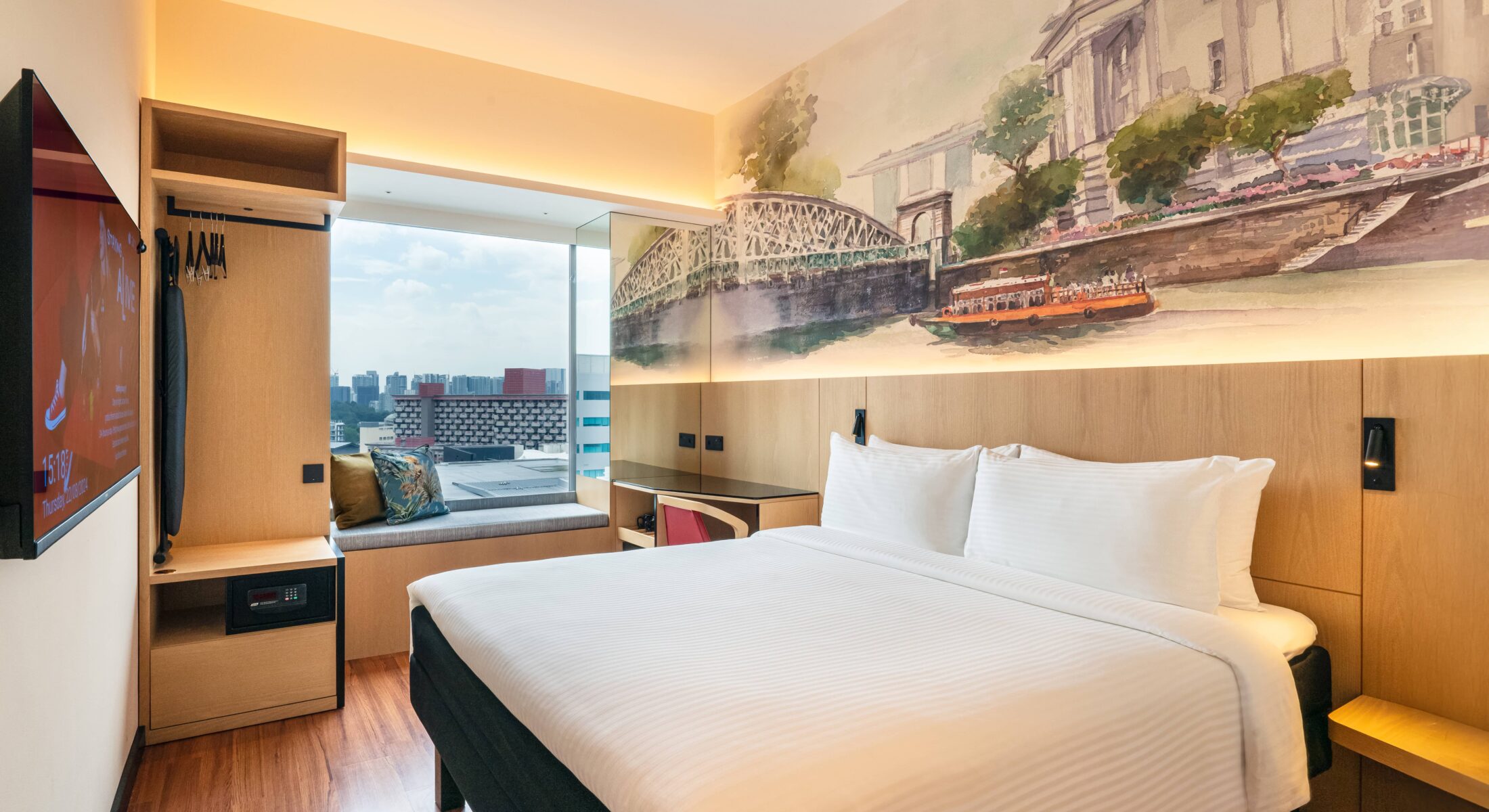 Spacious hotel room featuring a comfortable bed with a decorative headboard mural illustrating the Singapore River, a study table, and a bay window offering natural light.