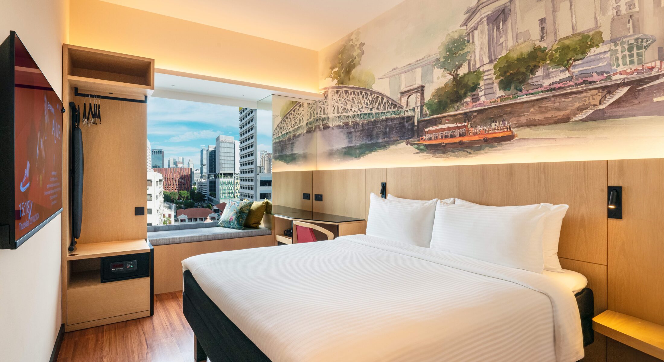 Spacious hotel room featuring a comfortable bed with a decorative headboard mural illustrating the Singapore River, a study table, and a bay window offering natural light.