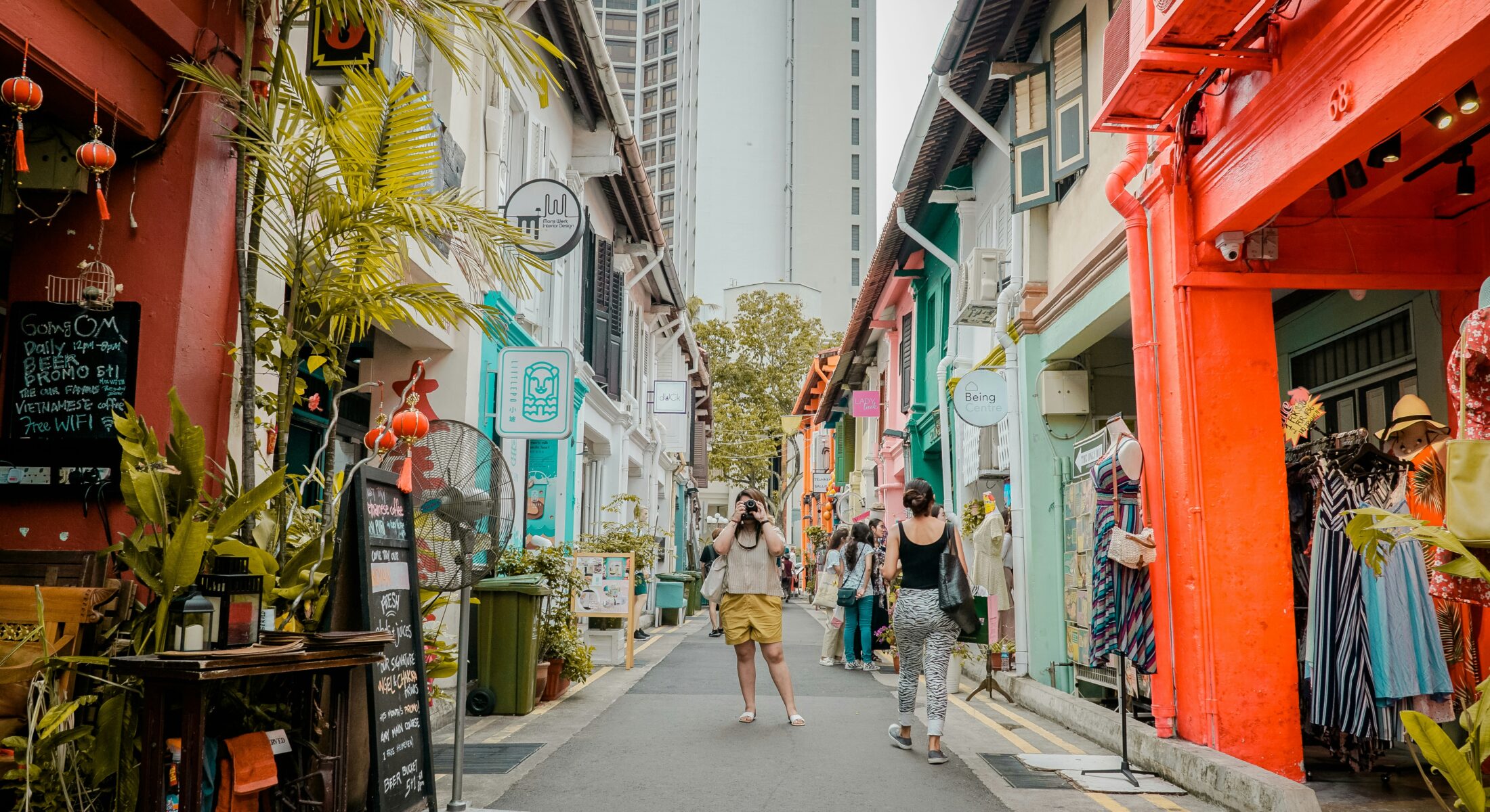 A vibrant street in Singapore with colorful shophouses, boutique stores, and cafés. A tourist in yellow shorts is taking a photo, while another person walks past. The street is lined with plants, signage, and traditional architecture.