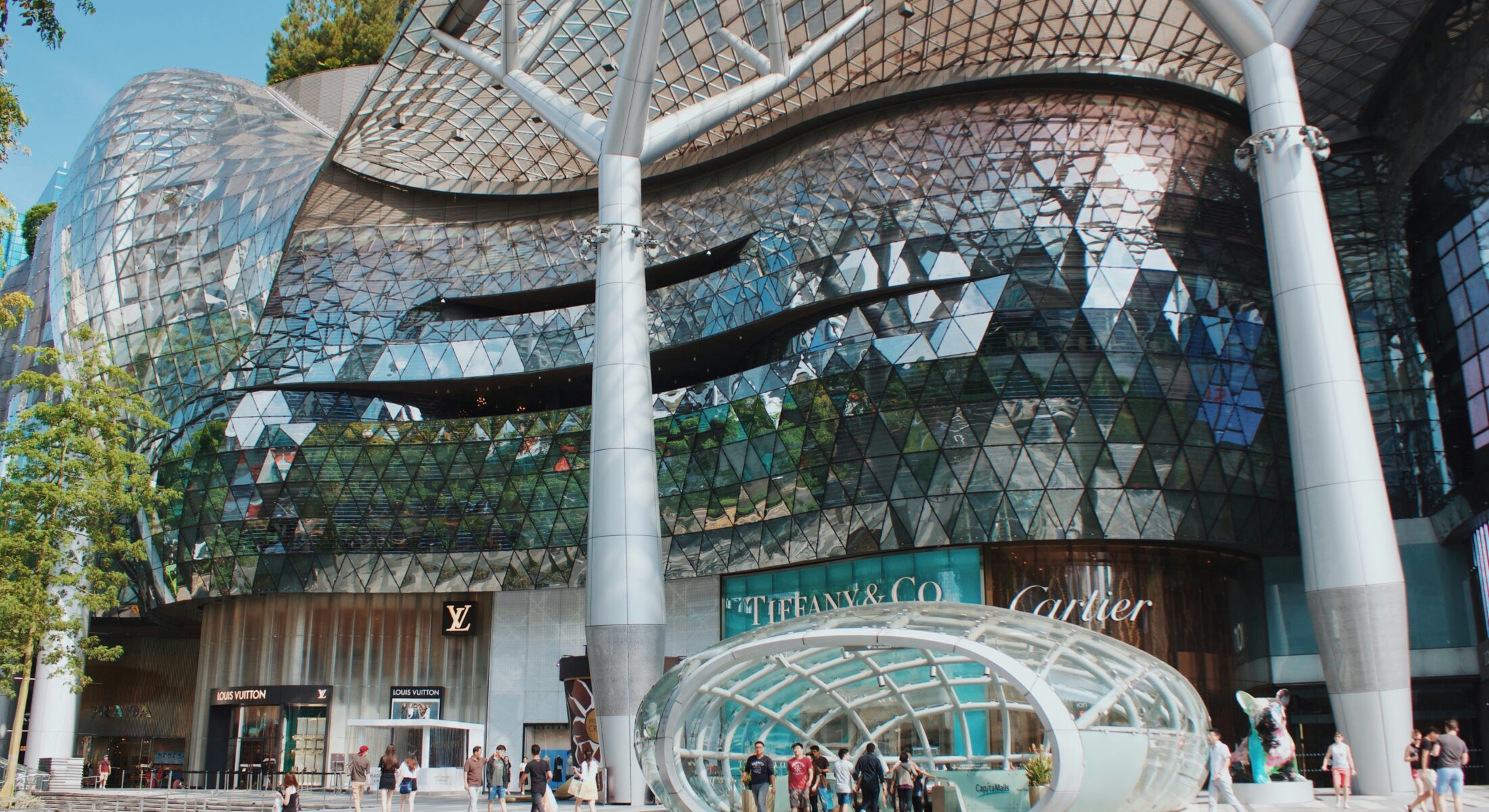 ION Orchard shopping mall in Singapore, featuring a futuristic glass façade with geometric patterns. Luxury brands like Louis Vuitton, Tiffany & Co., and Cartier are visible at the entrance, with people walking around the plaza.